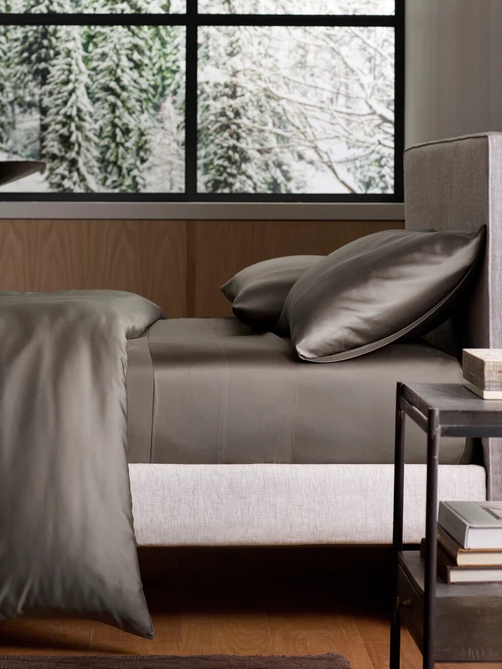 A modern bed with dark gray bedding and pillows next to a metal nightstand, with a snowy forest scene visible through the window behind.