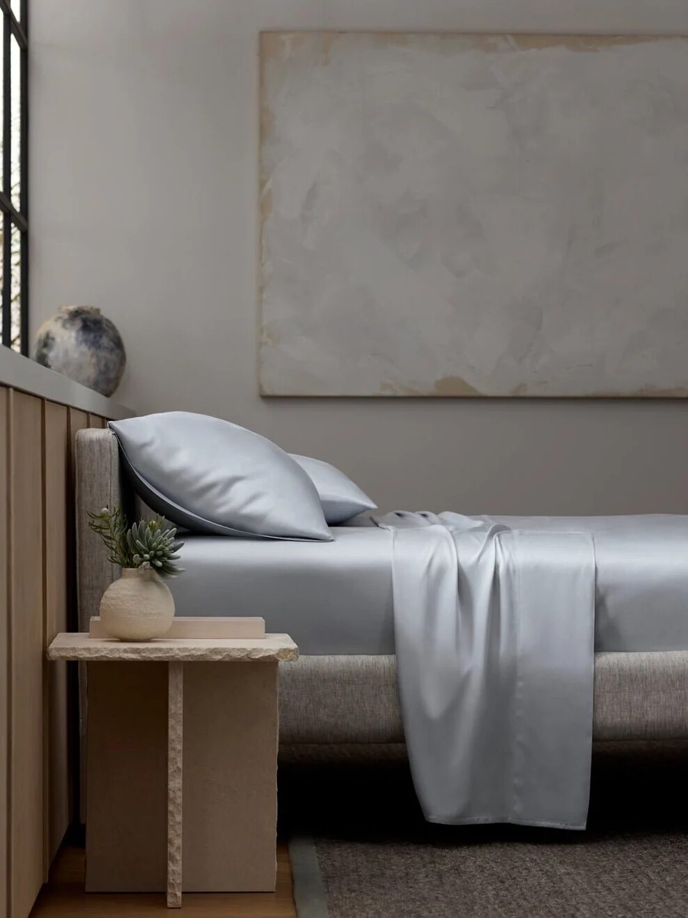 A neatly made bed with silver-gray satin sheets beside a small table holding a potted plant, in a minimalist bedroom with neutral decor and a large abstract wall art.