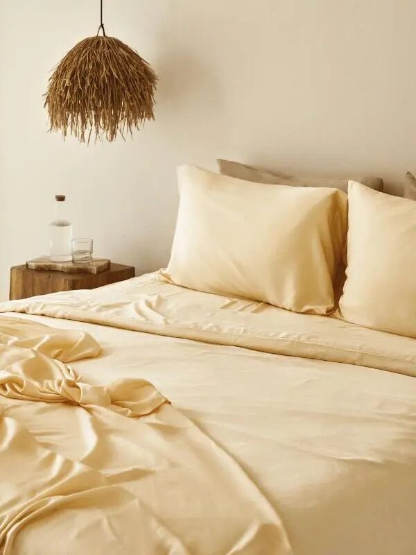 A neatly made bed with cream-colored sheets and pillows, a rustic hanging lamp, a wooden side table, and a glass water bottle by a sheer curtained window in a softly lit room.