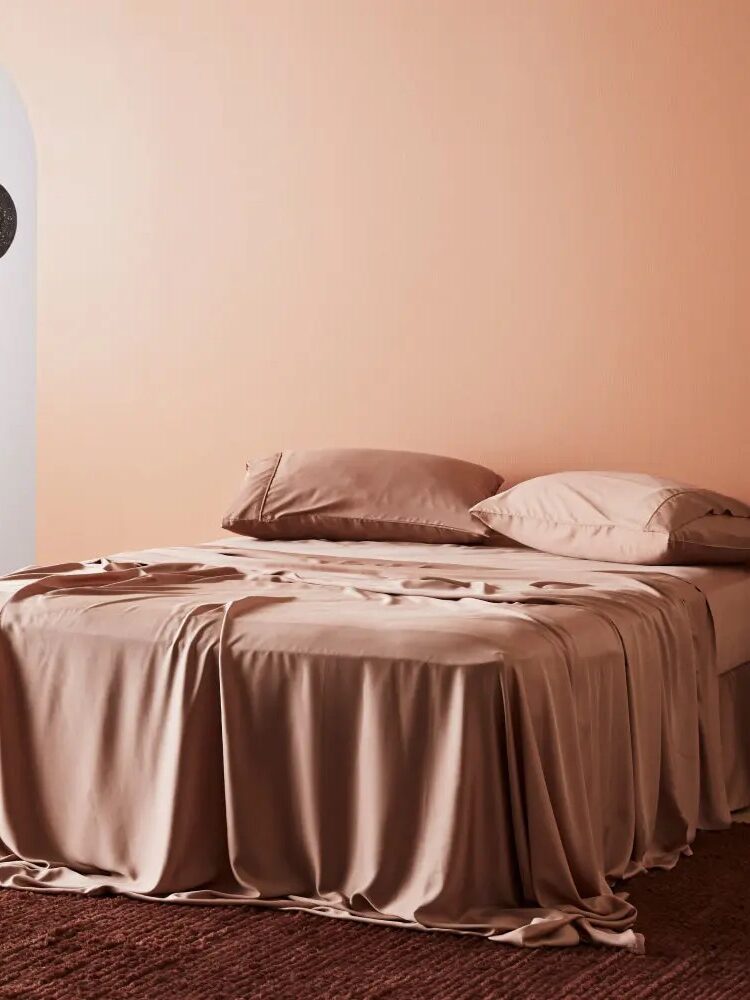 A neatly made bed with pink sheets and pillows sits on a textured rug in a pale peach room, with an arched doorway and a hanging decorative lamp in the background.