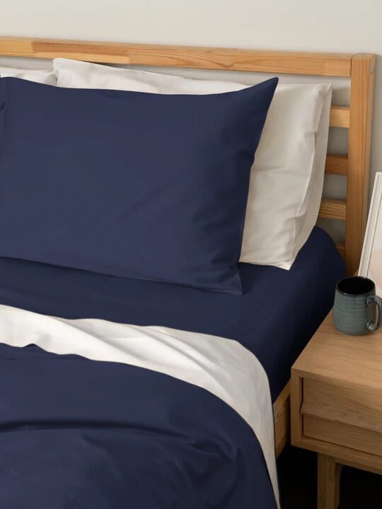 A neatly made bed with navy blue pillowcases and sheets, white bedding, and a light wooden headboard beside a nightstand holding a mug, book, and framed artwork.