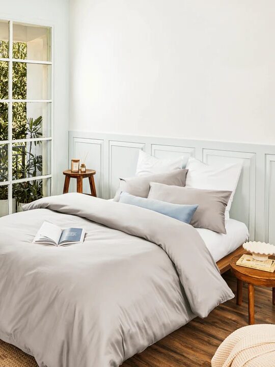 A neatly made bed with gray bedding and multiple pillows sits next to large windows with plants outside; an open book lies on the bed in a bright, modern bedroom.