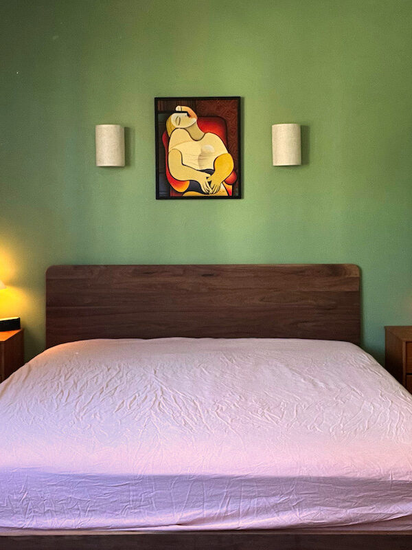 A neatly made bed with a wooden headboard, two bedside tables with lamps, and a framed abstract art piece on a green wall.