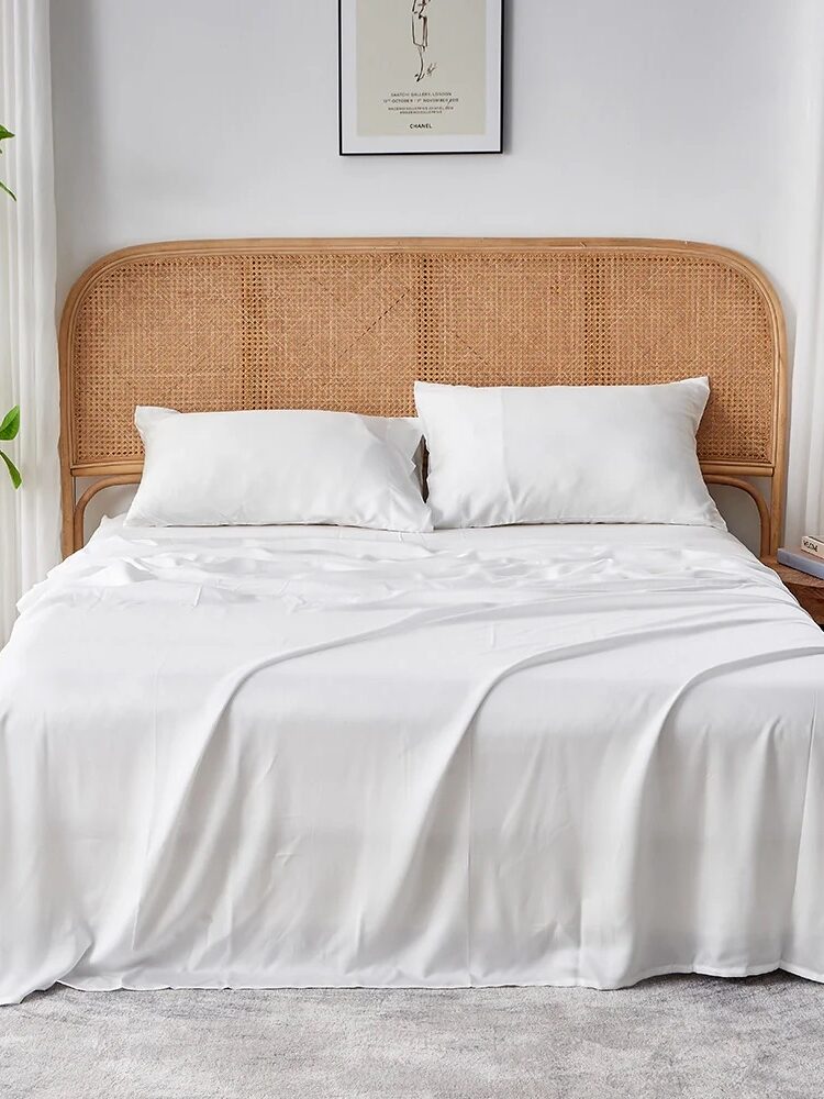 A neatly made bed with white sheets and pillows, a wicker headboard, bedside tables with a lamp and decor, a plant on the left, and artwork above the bed.
