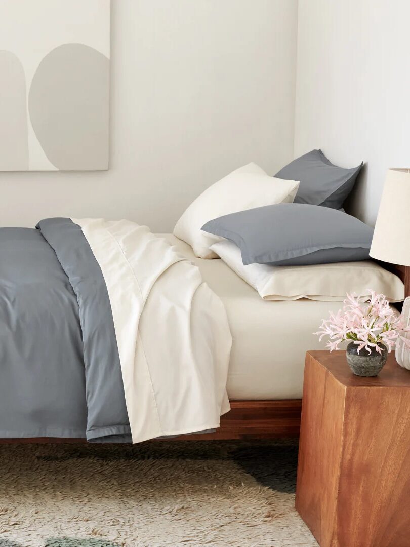 A neatly made bed with gray and white bedding, four pillows, a wooden nightstand with a lamp and pink flowers, and abstract art on the wall.