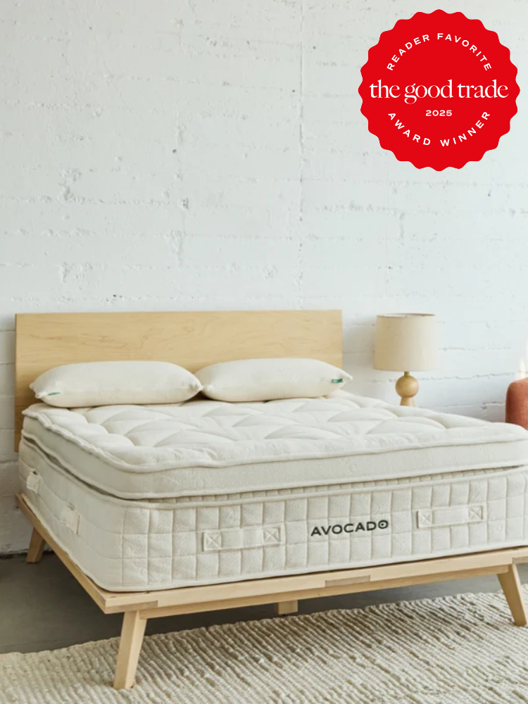 A minimalist bedroom with a wooden bed frame, an Avocado mattress, two pillows, a side table with a lamp, and a red "The Good Trade 2024 Award Winner" badge in the corner.