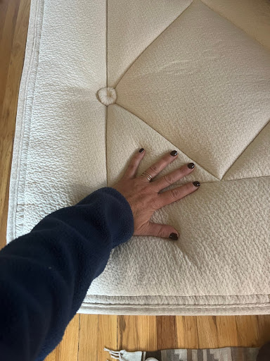A hand with dark nail polish presses down on the surface of a light-colored, textured mattress on a wooden floor.