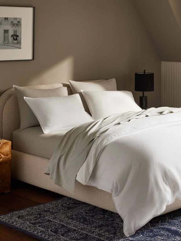 A neatly made bed with white bedding and four pillows sits on a blue patterned rug in a softly lit bedroom with beige walls and framed artwork.