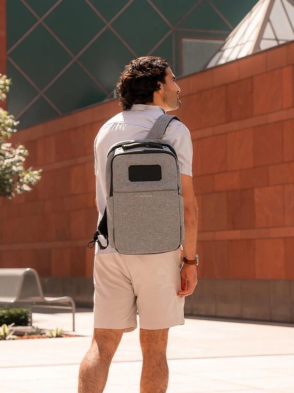 A person wearing a grey backpack and light-colored shorts stands outdoors in front of a modern building with a red and green facade.