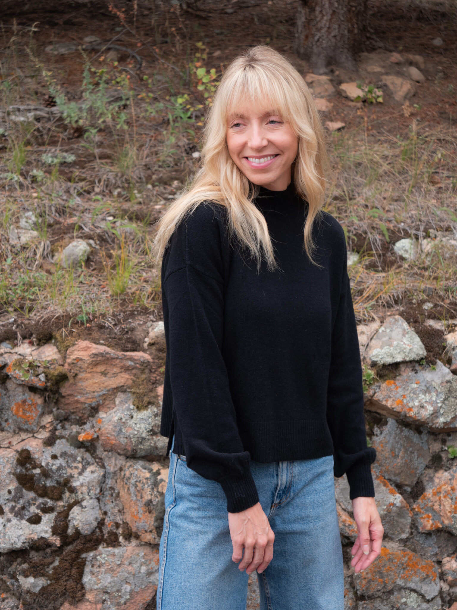 A woman with long blonde hair, wearing a black sweater and blue jeans, stands outdoors in front of a stone wall and grassy hillside, smiling slightly.