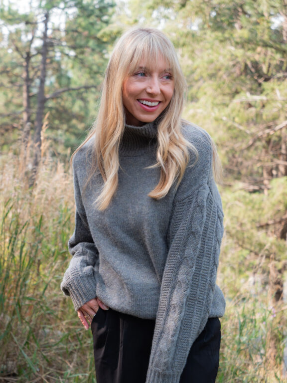 A woman with long blonde hair wearing a gray knit sweater and black pants stands outdoors in a forested area, smiling slightly and looking to the side.