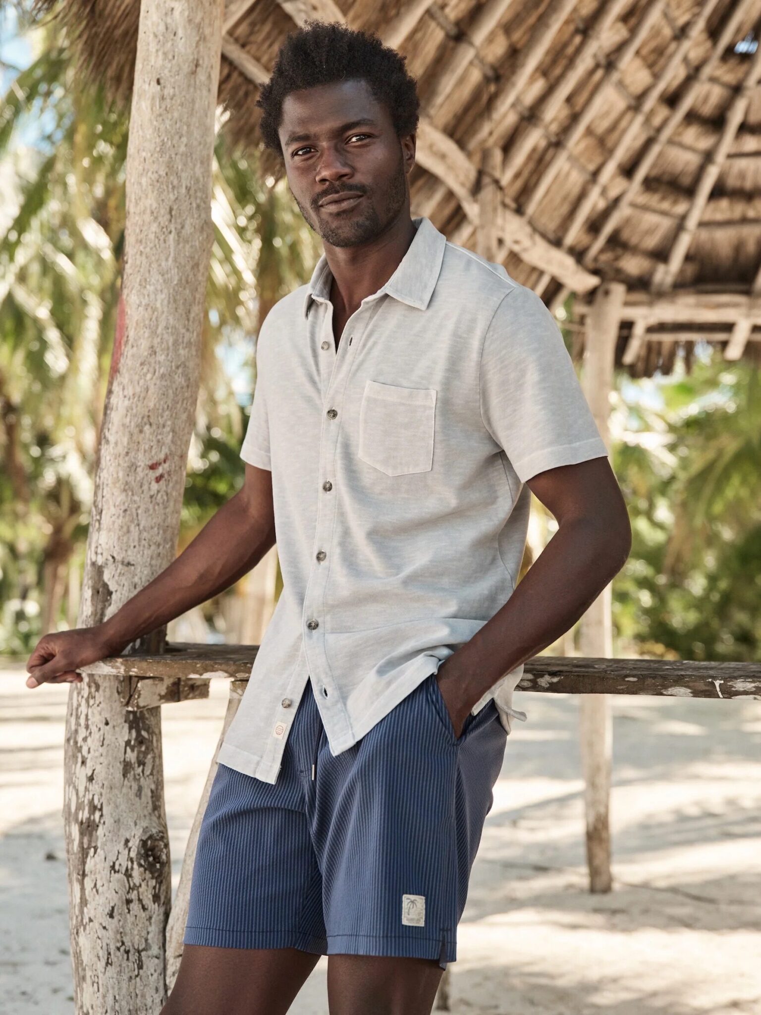 A man stands by a wooden structure on a sandy beach, wearing a light short-sleeve shirt and blue shorts, with palm trees in the background.