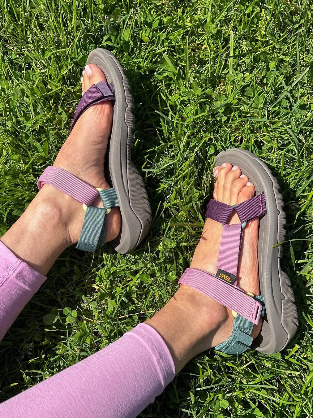 Person wearing colorful Teva sandals and light purple leggings, sitting on green grass.