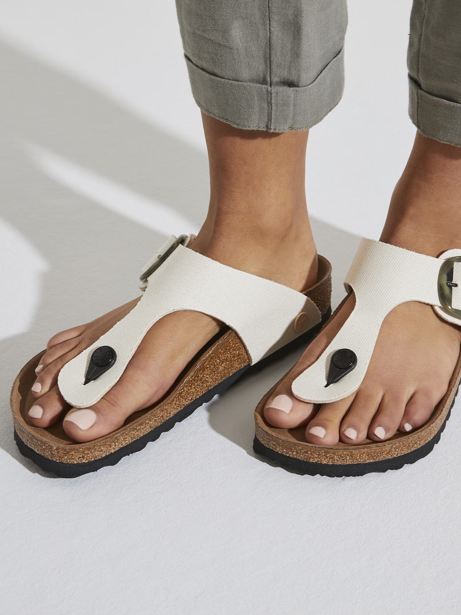 A person wearing light-colored sandals with cork soles and black buckles, standing on a white surface with rolled-up gray pants.