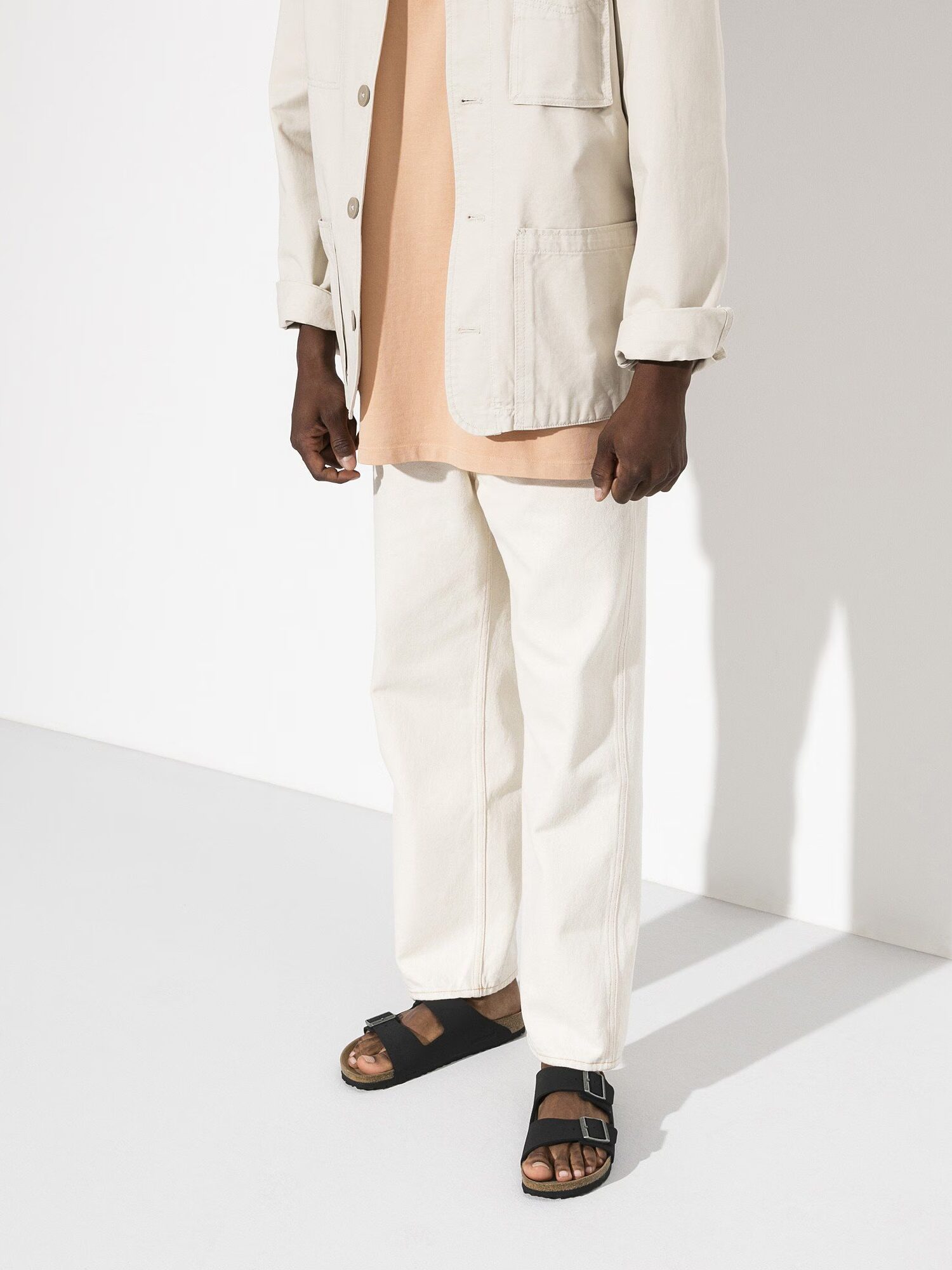A person dressed in a beige jacket, peach shirt, off-white pants, and black sandals standing against a white background.