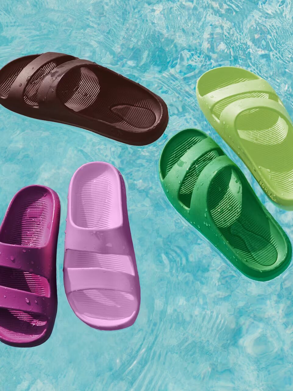Four pairs of slide sandals in brown, green, purple, and pink float on the surface of clear blue pool water.