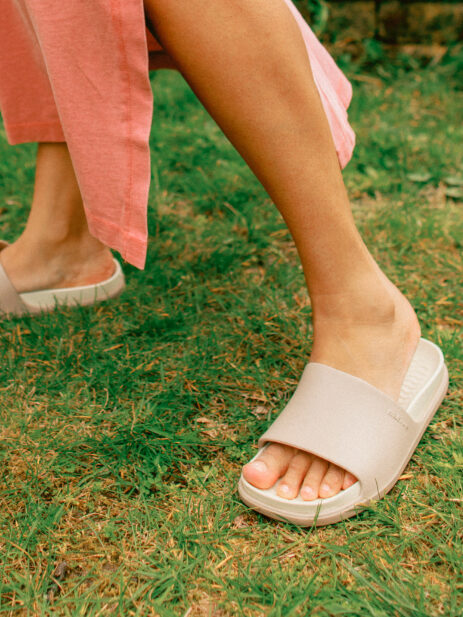 A person wearing light beige slide sandals and a pink skirt walks on green grass.