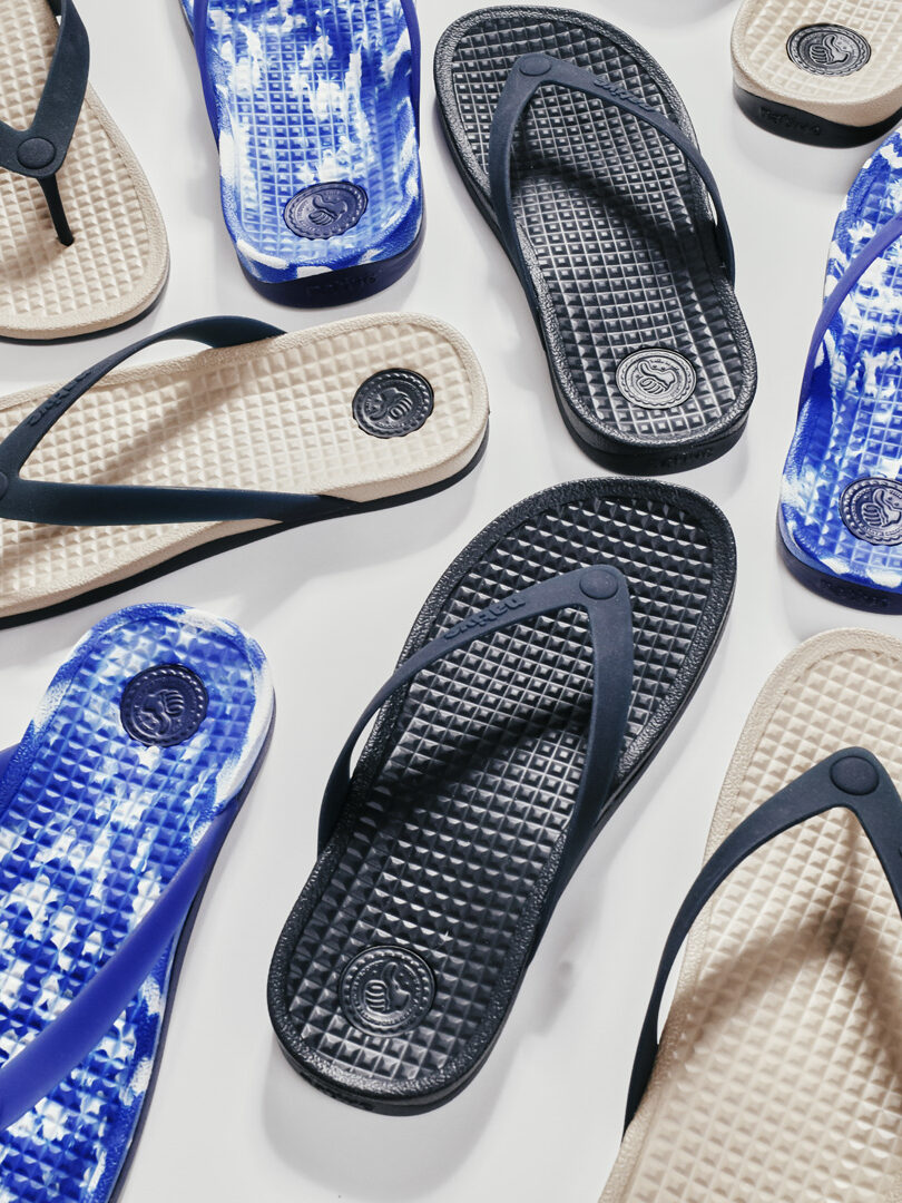 Several pairs of flip-flops are arranged on a white surface, featuring solid black, solid beige, and blue-and-white tie-dye patterns.