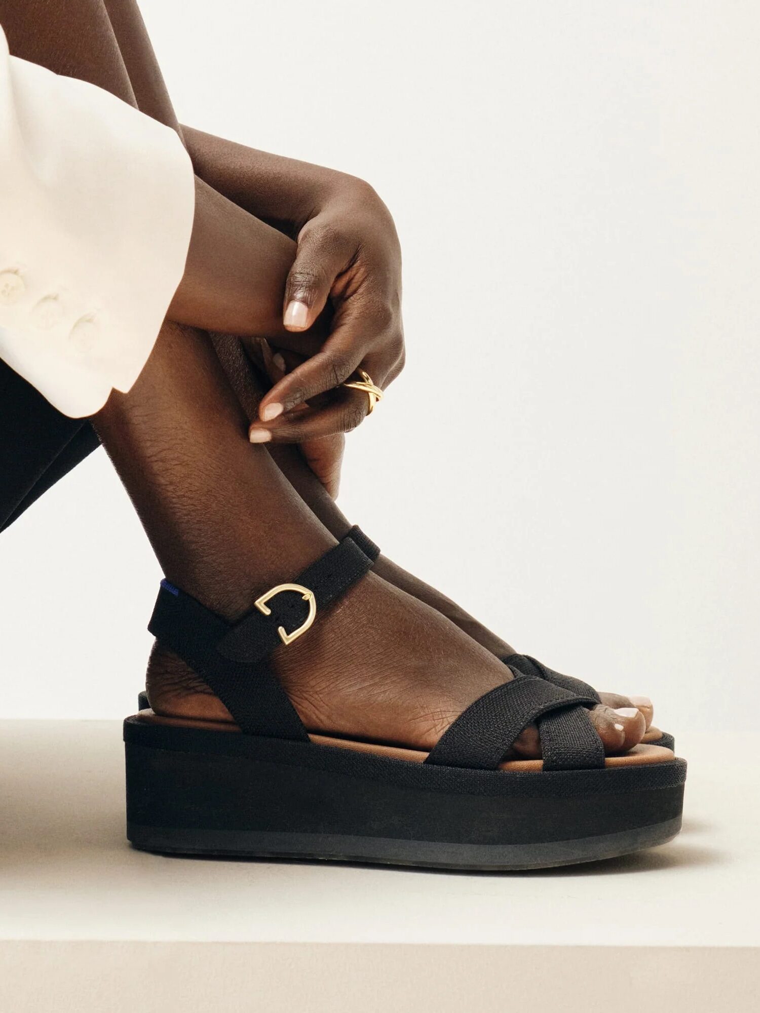 A person in a white blazer and black pants sits with one leg crossed, showing black platform sandals with gold buckle accents.