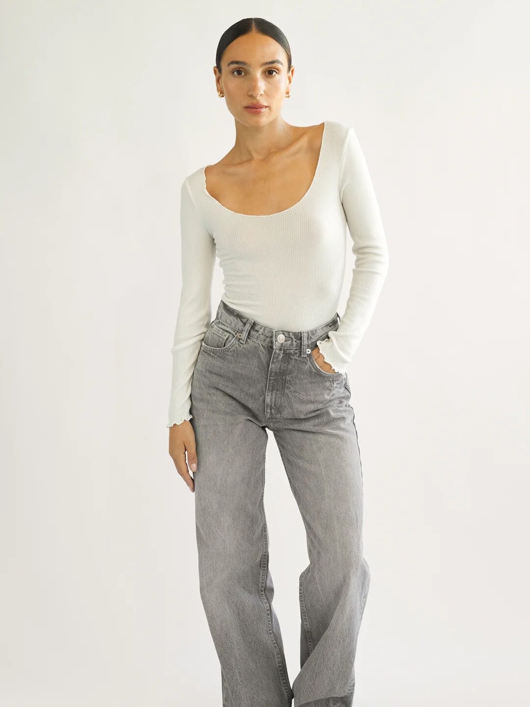A person stands against a plain background wearing a white long-sleeve top, gray high-waisted jeans, and black sandals.