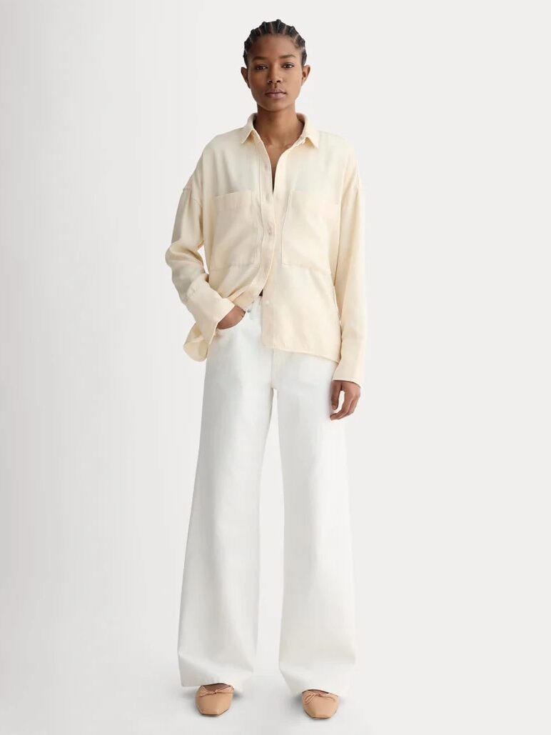 A person stands facing forward wearing a light beige button-up shirt, wide-leg white pants, and beige shoes against a plain light background.