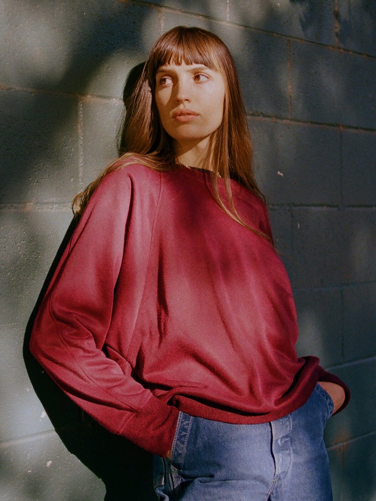 A person with long brown hair wearing a red sweatshirt and blue jeans stands against a sunlit brick wall with hands in pockets.