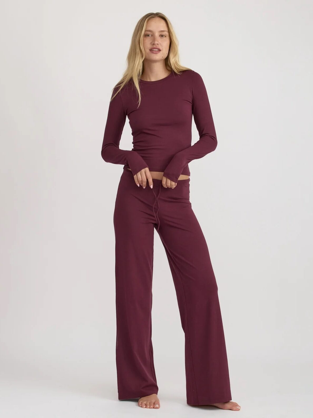 A woman stands barefoot against a plain background, wearing a long-sleeve, burgundy top and matching wide-leg pants.
