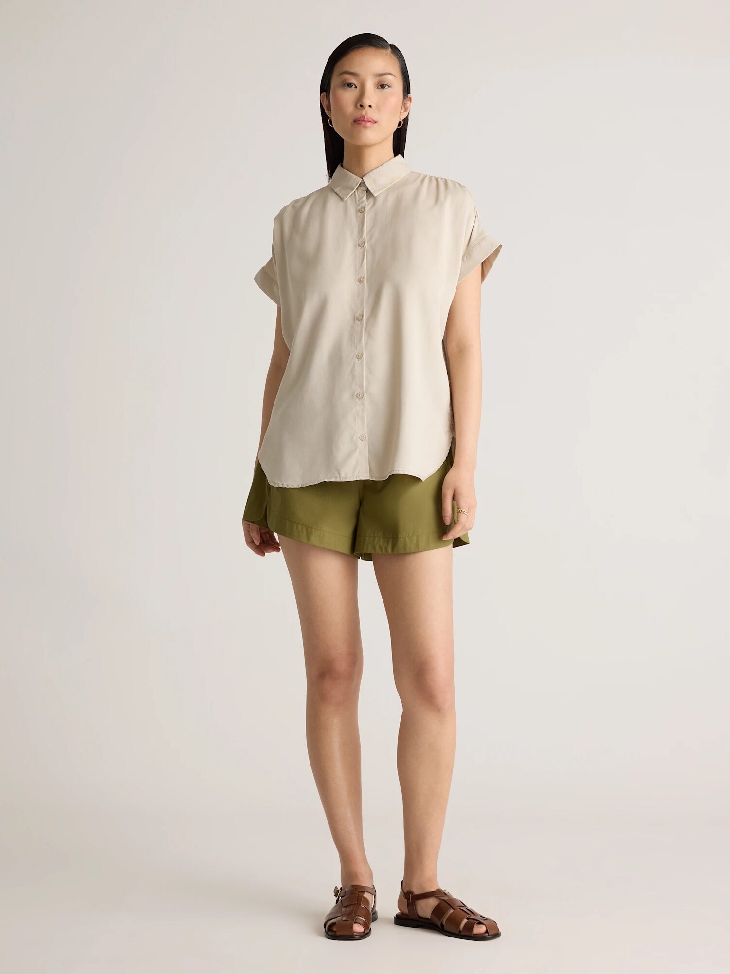 A person stands facing forward wearing a short-sleeve beige button-up shirt, olive green shorts, and brown sandals against a plain background.