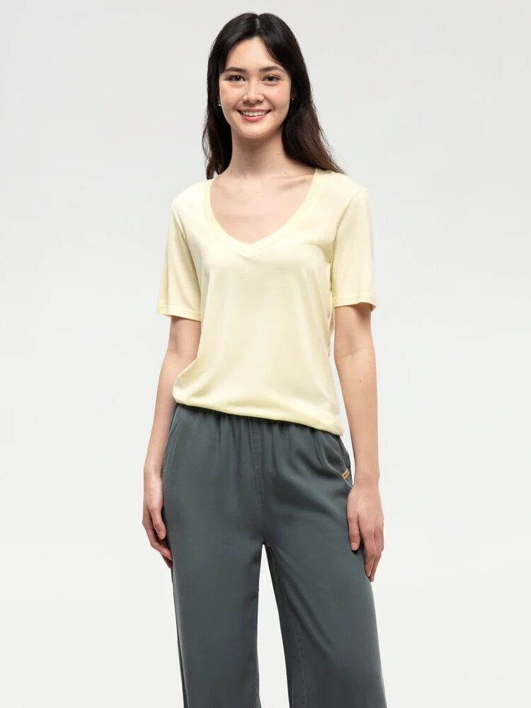 A woman stands smiling, wearing a light yellow short-sleeve shirt and dark gray pants against a plain light background.