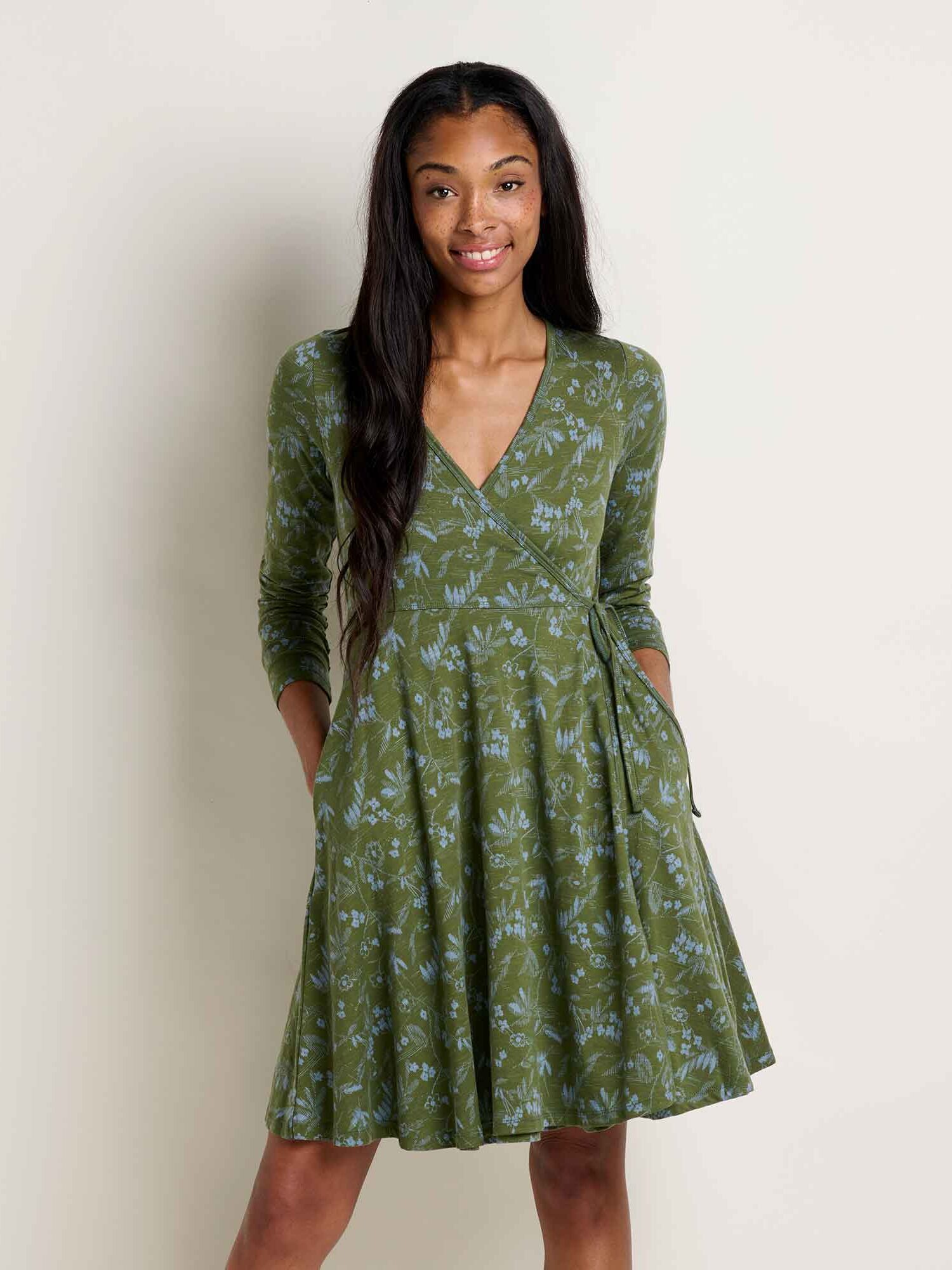 A woman stands against a plain background wearing a long-sleeve, green wrap dress with a subtle light blue floral pattern and pockets.