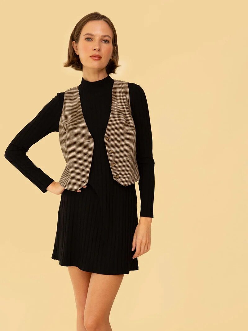 A person stands against a beige background wearing a black long-sleeve dress and a brown houndstooth vest, with one hand resting on their hip.