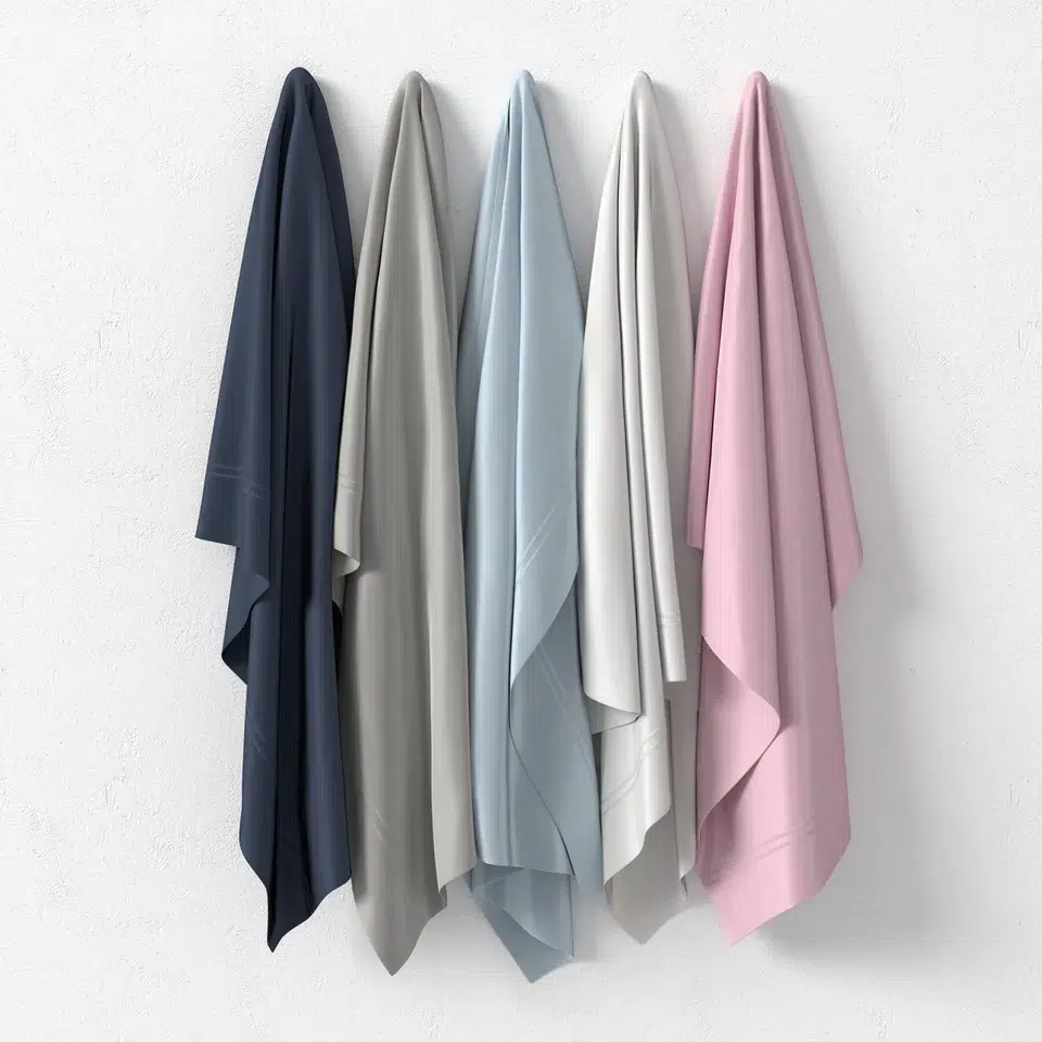 Four towels in navy blue, light gray, light blue, and pink are hanging side by side on hooks against a white wall.
