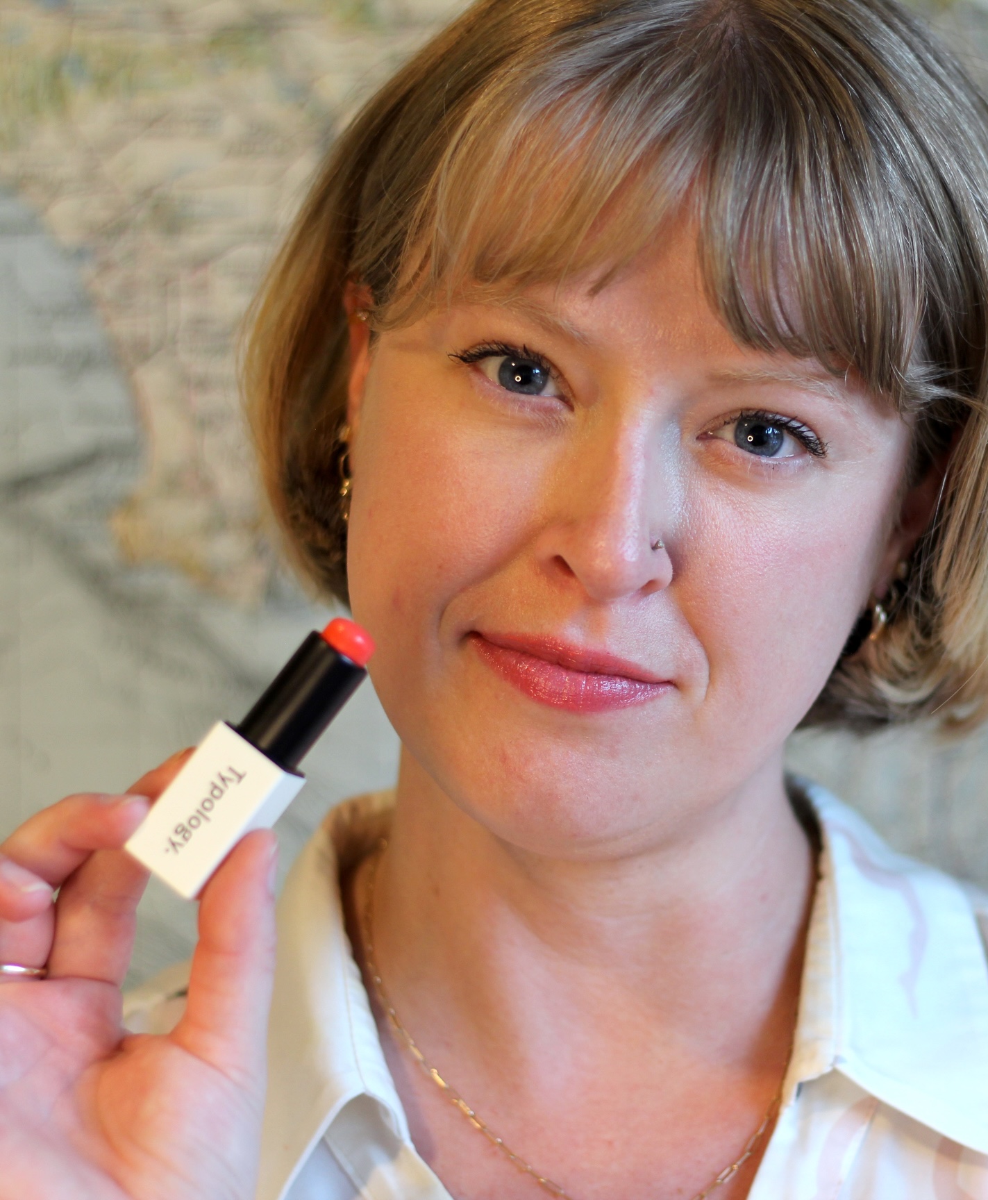 A woman with short blonde hair holds a tube of lipstick near her face, wearing a white shirt and standing in front of a blurred map background.