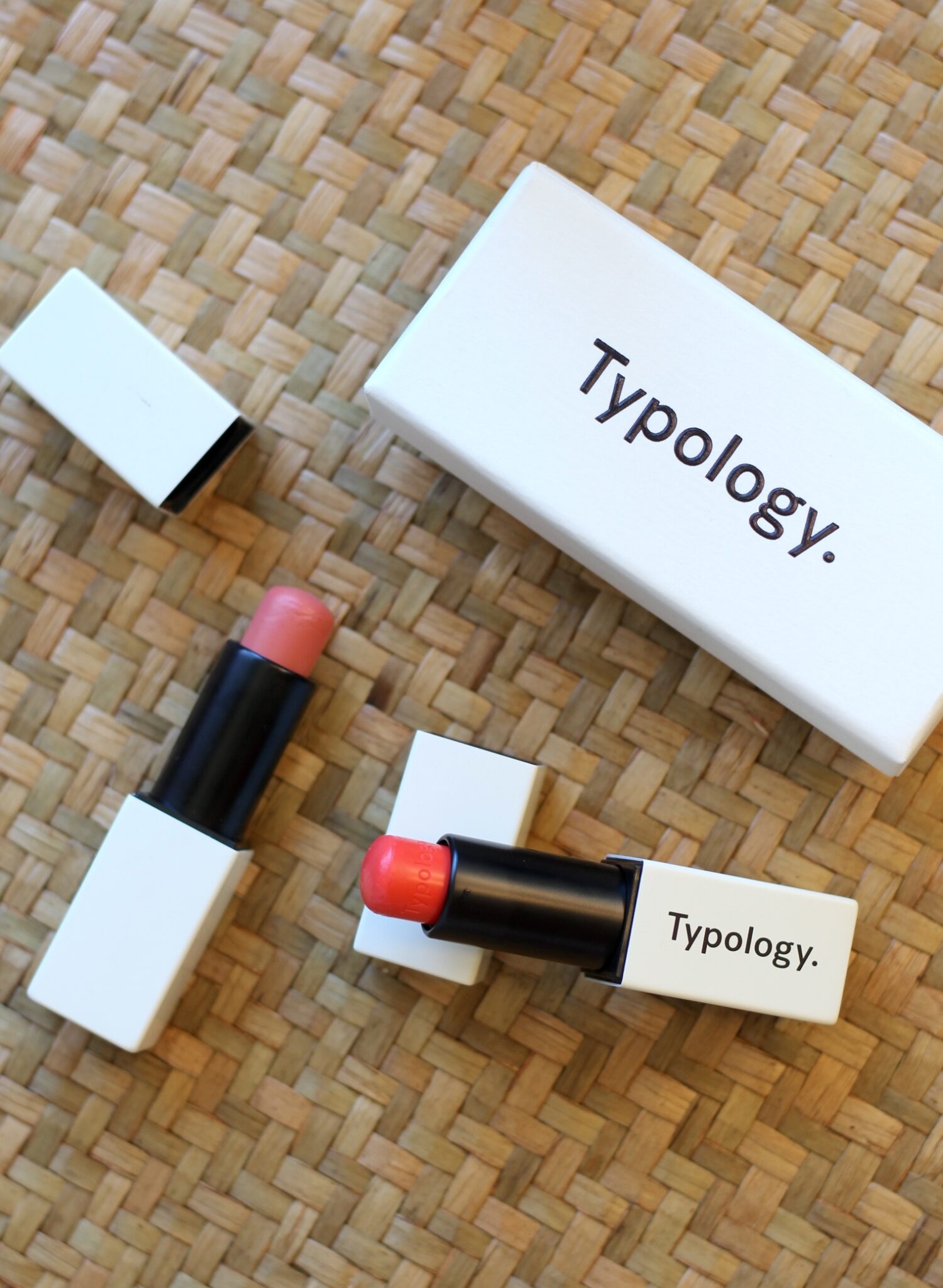 Two Typology lipstick tubes, one peach and one red, with their caps off, are placed on a woven mat next to a branded Typology box.