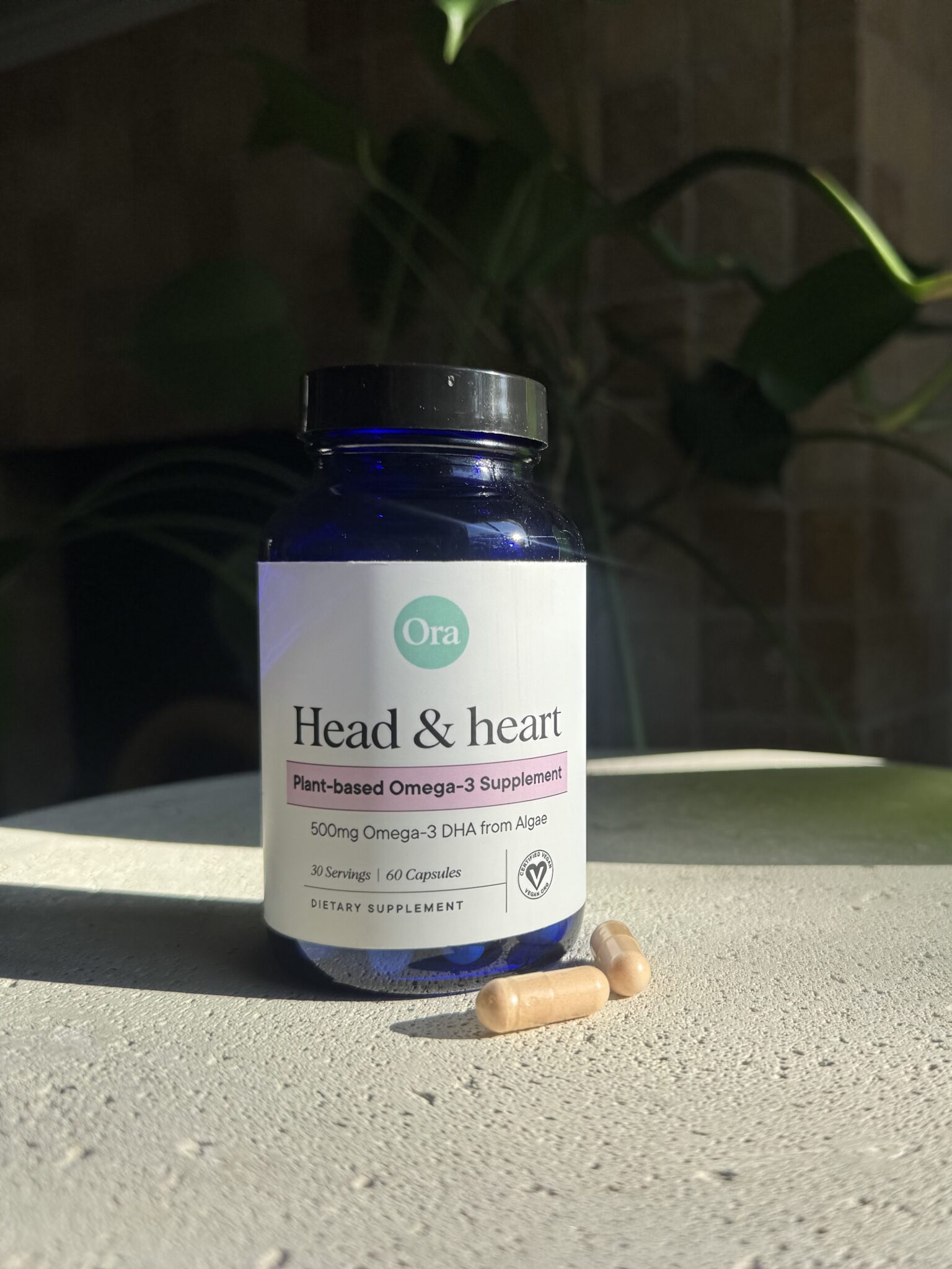 A bottle of Ora Head & Heart plant-based Omega-3 supplement sits on a surface in sunlight, with two capsules in front of it and green plant leaves in the background.