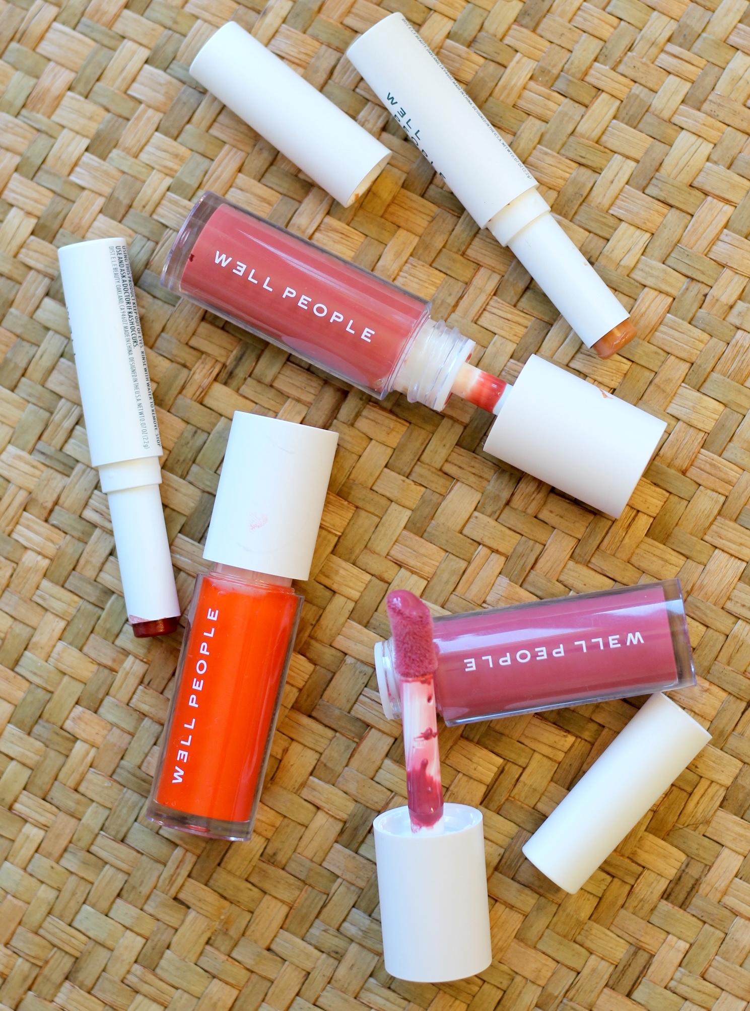 Four WELL PEOPLE lip products, including two open lip gloss tubes and two lip crayons, are arranged on a woven surface.
