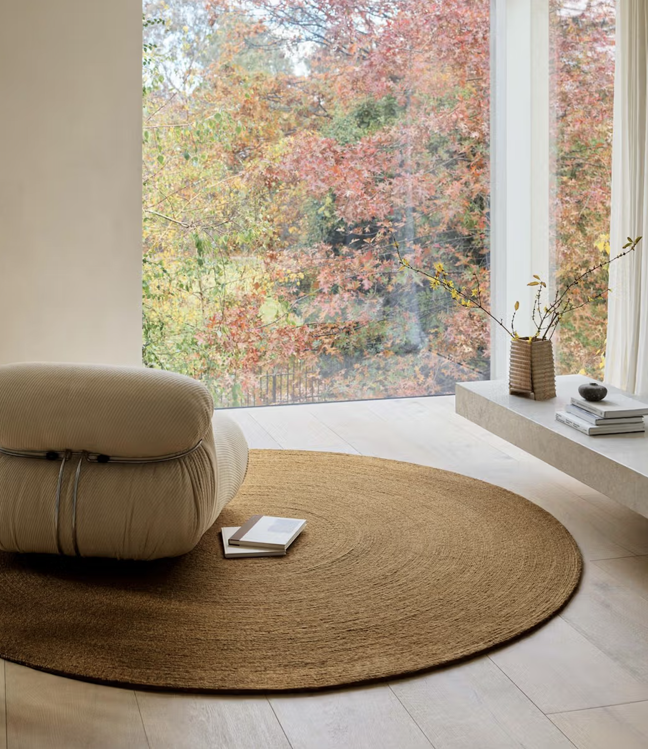 A beige lounge chair and a round woven rug are placed near a large window with a view of autumn trees. A low shelf holds a vase with branches, a bowl, and a stack of books.