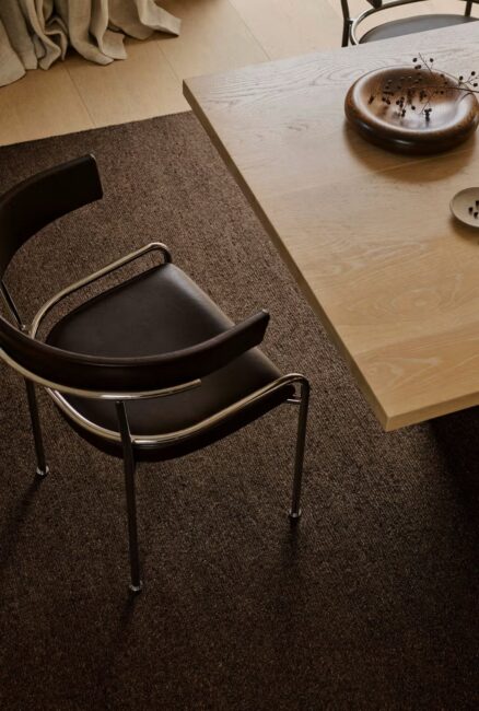 A black chair with a metal frame sits next to a light wood table with a round wooden bowl and a small plate on a brown carpeted floor.
