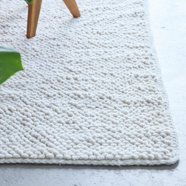 Close-up of a textured white woven rug with small round patterns on a light-gray floor, partially showing a wooden chair leg and green plant leaf in the corner.