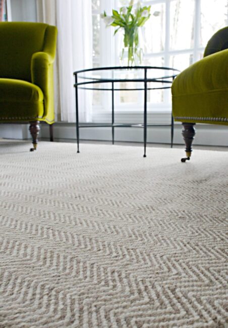 Two green armchairs and a glass-top round table are placed on a textured cream carpet in front of a large window with sheer curtains.