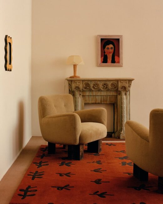 Two beige armchairs face each other on a red patterned rug in front of a decorative mantel with a lamp and framed portrait on the wall above.