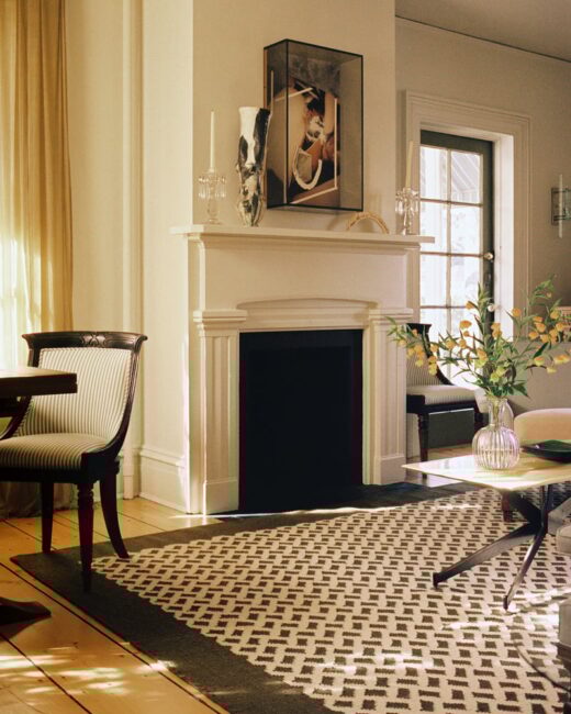 A living room with a black fireplace, a patterned rug, a vase with yellow branches on a coffee table, and framed art above the mantel. Sunlight streams through tall windows with sheer curtains.