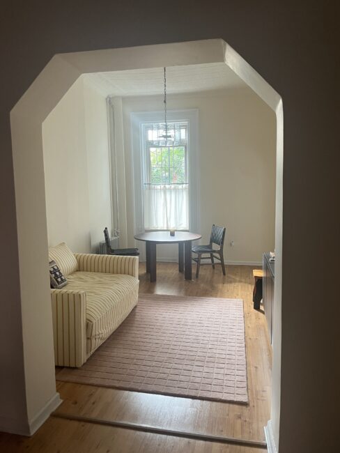 A small room with a striped sofa, a round table with two chairs, a pink rug, and a window with sheer curtains letting in natural light.