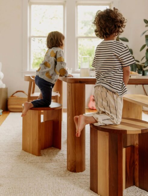 Two young children sit on wooden stools at a wooden table in a bright, sunlit room with large windows and a white rug.