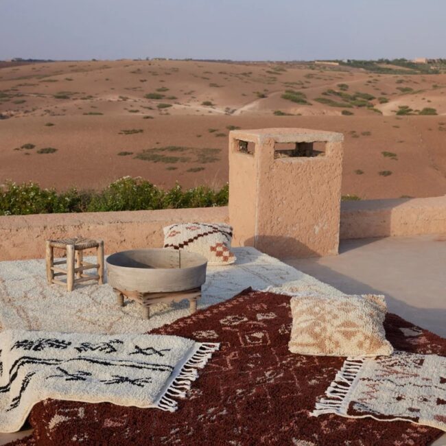 Outdoor patio with patterned rugs, pillows, a small wooden stool, and a round wooden basin, overlooking a desert landscape with sparse greenery.