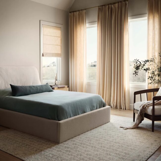 A modern bedroom with a large bed, light blue bedding, beige rug, armchair, potted plant, wooden side table, and floor-to-ceiling curtains covering two windows.