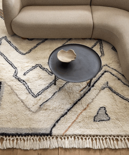A curved beige sofa sits on a patterned cream rug with black lines, next to a round black coffee table holding a beige ceramic bowl.
