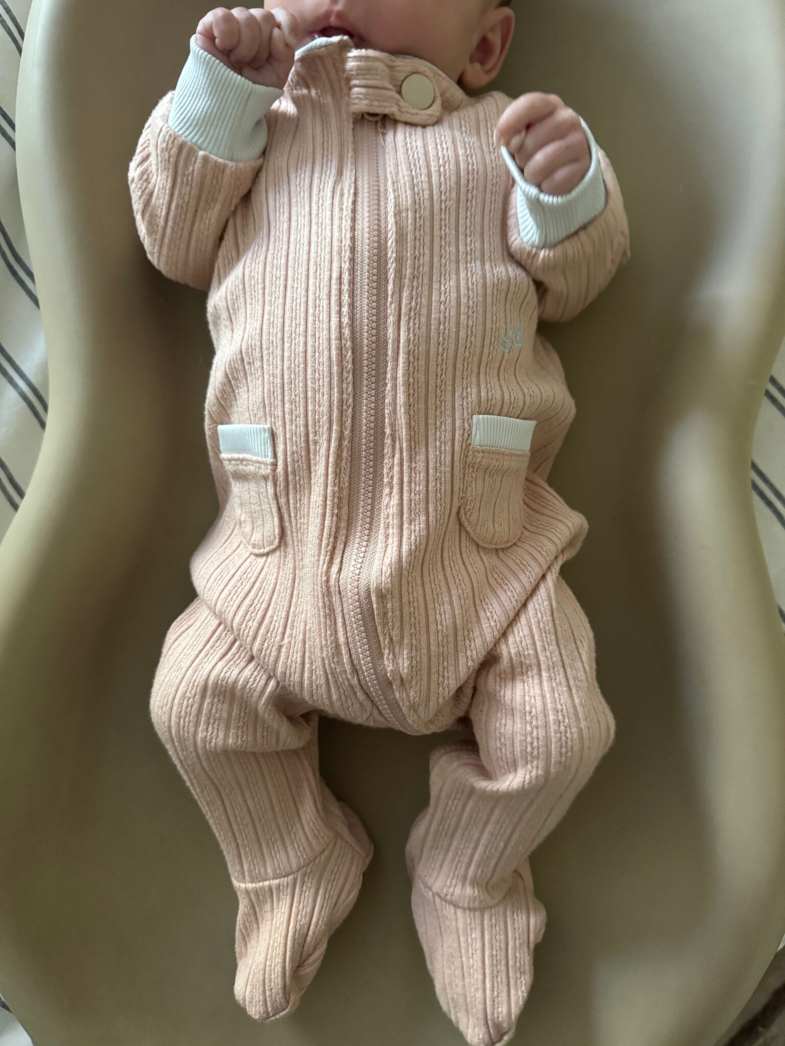 Baby in a light pink ribbed onesie with white cuffs lying in a beige seat, holding a hand near their mouth.