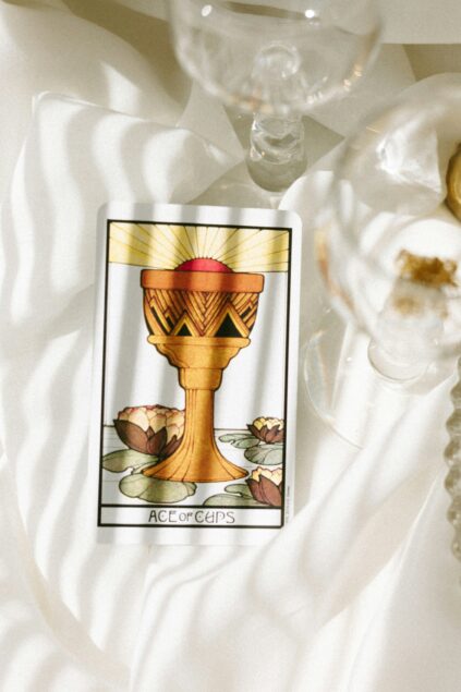 A tarot card labeled "Ace of Cups" featuring a golden chalice with water lilies, placed on a white fabric surface next to a glass.