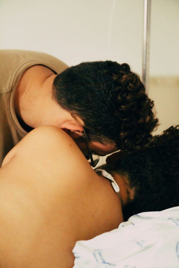 A person leans over and embraces another person lying in a hospital bed, wearing a medical gown and an IV line.